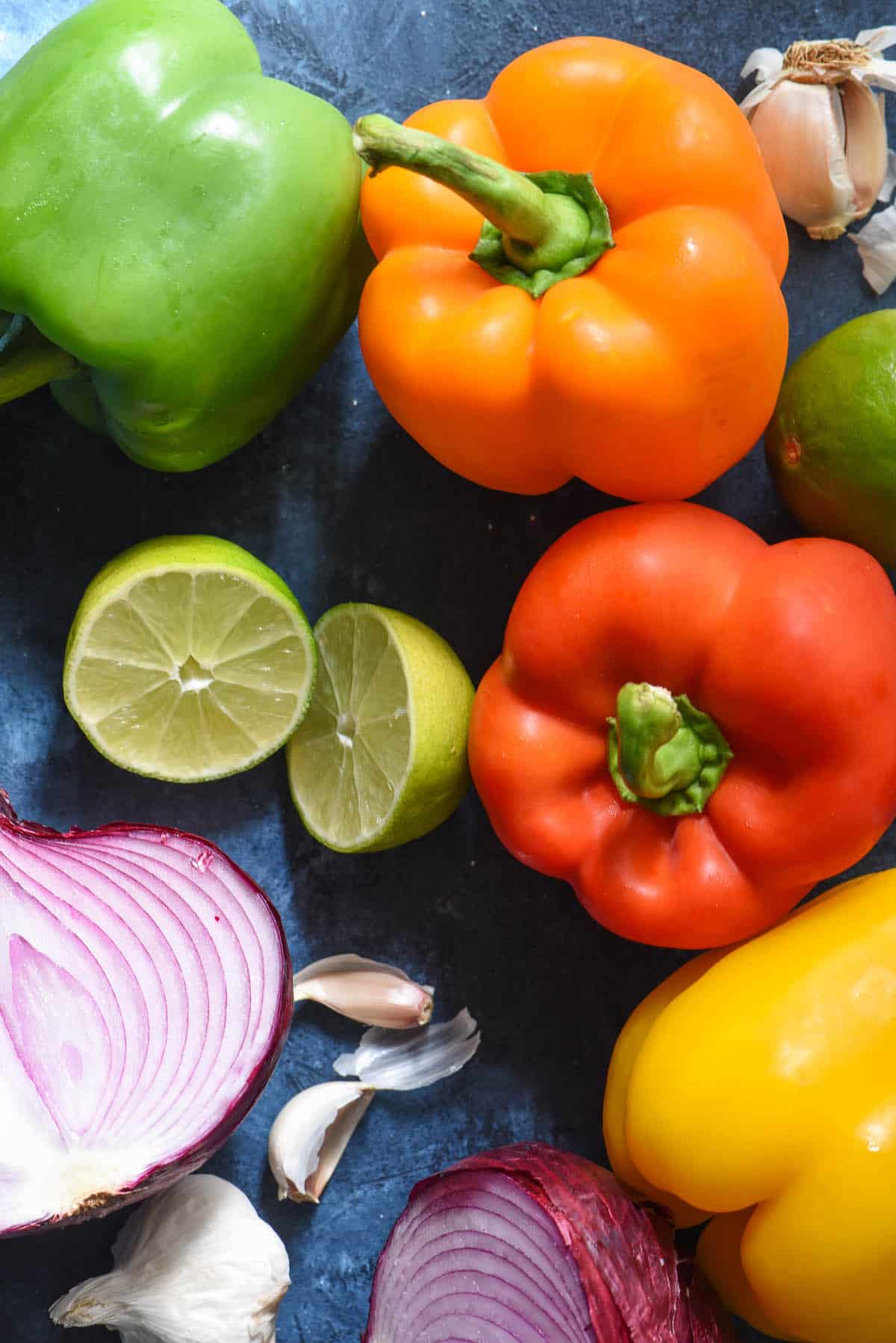 These Fajita Veggies have the restaurant flavor you love, made at home without a seasoning packet. Use them for vegetarian fajitas, or add steak, chicken or shrimp to the mix! | foxeslovelemons.com Raw bell peppers, lime, garlic and red onion on blue background.
