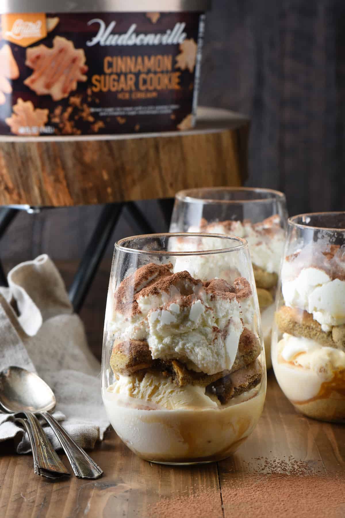 These Frozen Tiramisu Cups are a decadent five minute treat in a glass! Layers of coffee-soaked lady fingers, ice cream and mascarpone whipped cream. | foxeslovelemons.com Tiramisiu trifles in stemless wine glasses, with Hudsonville Cinnamon Sugar Cookie ice cream carton on stand in background.
