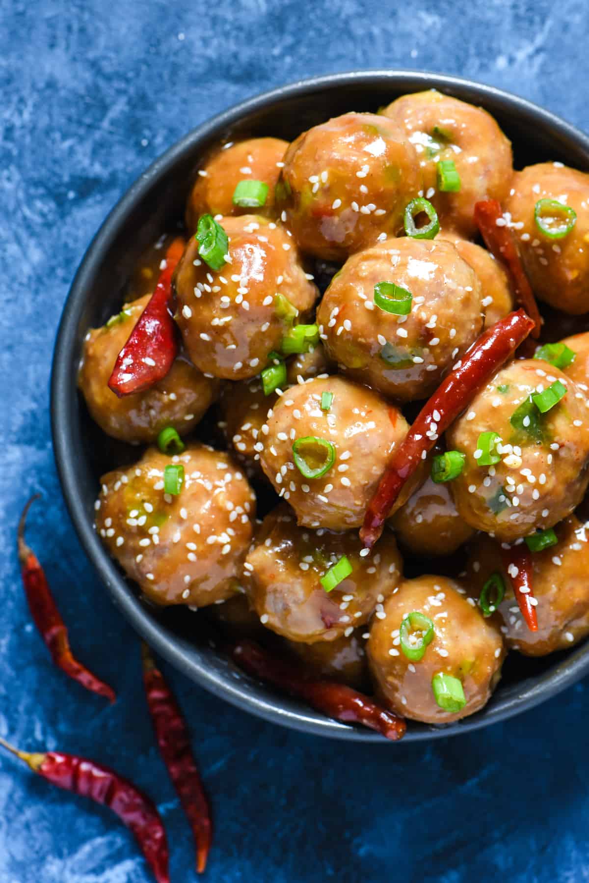 Round bowl of General Tso's meatballs topped with green onions, sesame seeds and dried chiles on a blue background.