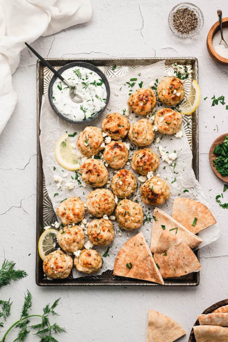 Mediterranean meatballs on a rimmed baking pan with pita wedges, lemon slices and yogurt sauce.