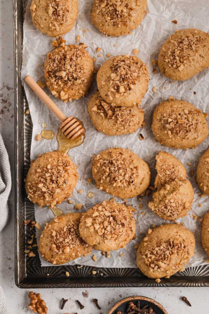 Melomakarona sprinkled with walnuts on white parchment paper with a honey dipper.