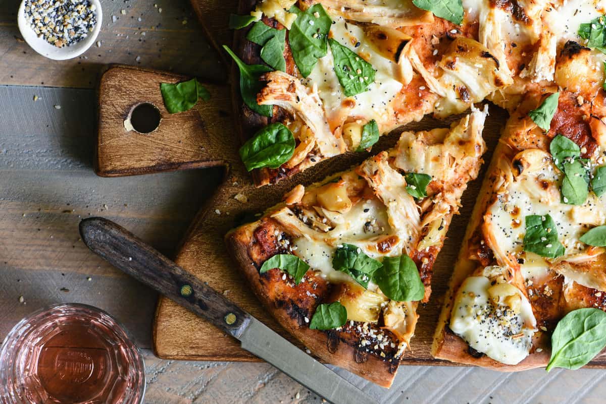 If you love homemade pizza, you have to try this Chicken Pizza with Mozzarella & Roasted Garlic! This recipe provides step-by-step instructions to either grill it like a pro, or make it in your oven. | foxeslovelemons.com Overhead photo of pizza with chicken, roasted garlic and greens, on cutting board with knife.