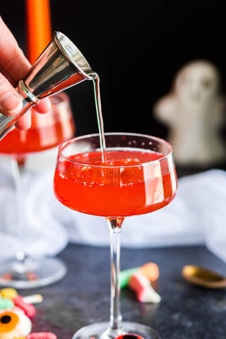 A hand pouring a shot of clear liquor into a coupe glass filled with a red drink.