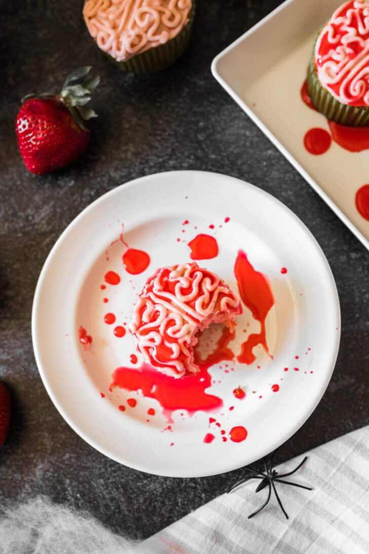 A Halloween cupcake piped with frosting to look like a bloody zombie brain, on a white plate, with a bite taken out of it.