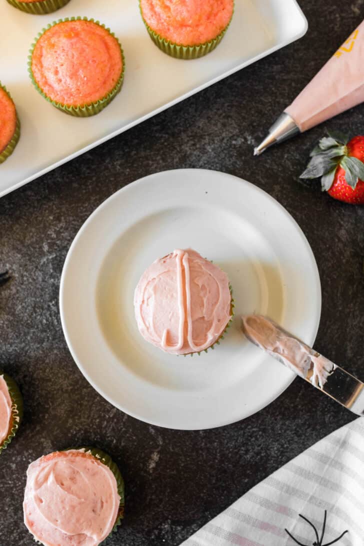 A cupcake on a plate, frosted with light pink frosting, with the start of a zombie brain design being piped on top.