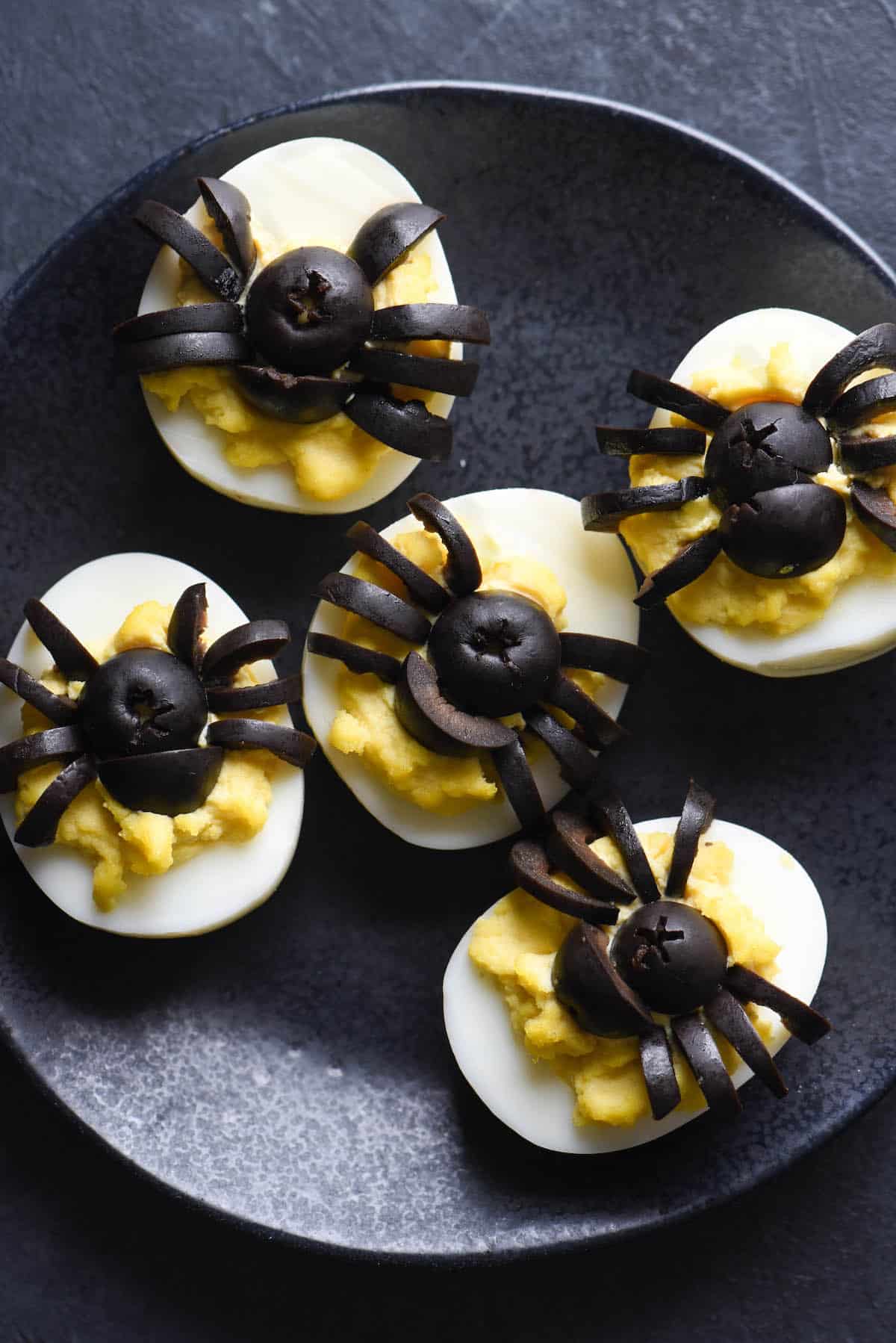 These Halloween Deviled Eggs are a spooky treat. The "trick" is that this Halloween party snack is also super easy and fast to put together! | foxeslovelemons.com Black plate with spider deviled eggs on top. Black olives are cut into the shape of spiders to decorate the tops of the eggs.