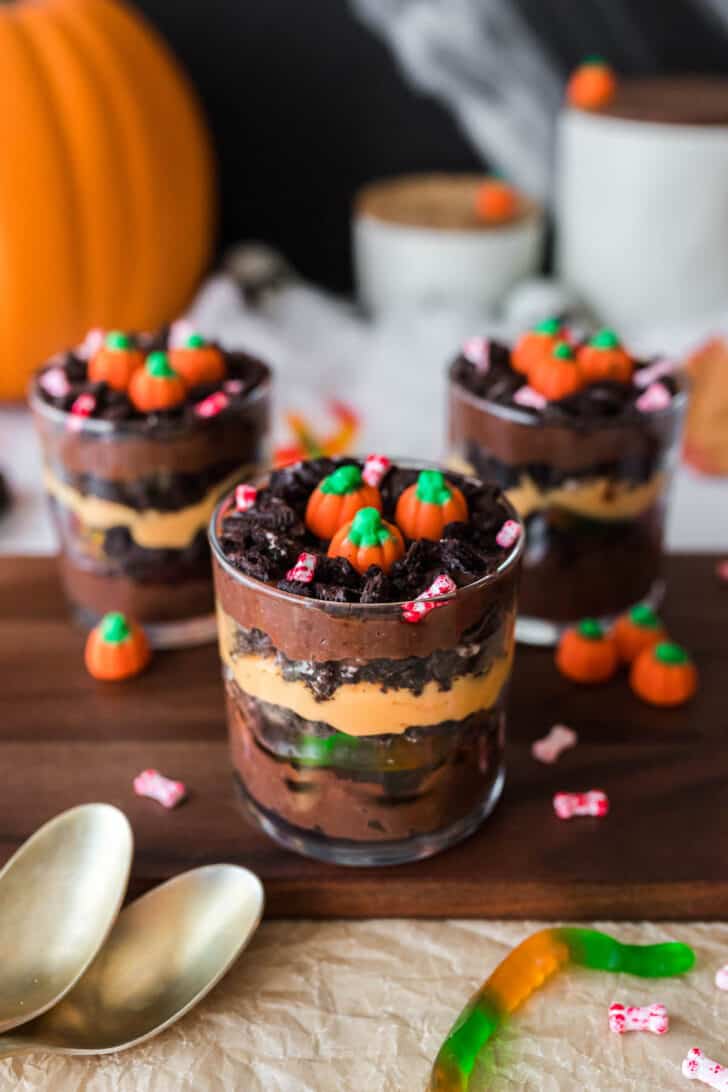 Halloween dirt cups made with layers of chocolate and orange colored pudding, crushed cookies and gummy worms. Three cups are arranged on a wooden cutting board and topped with candy pumpkins.