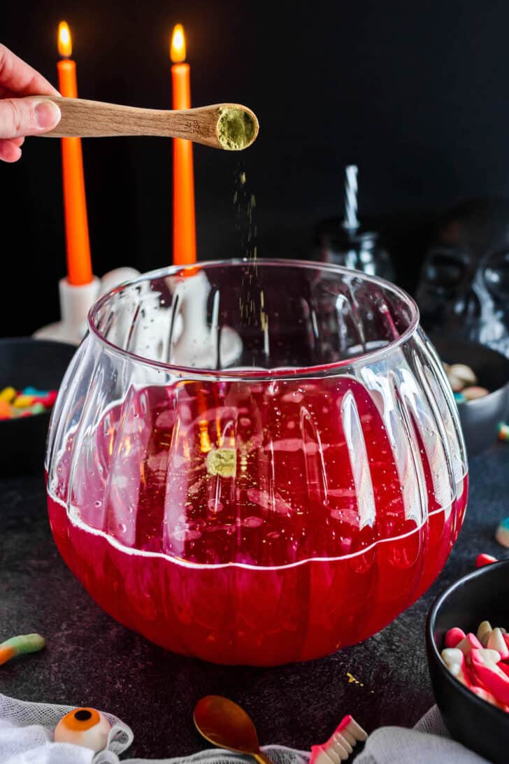 A hand sprinkling a spoonful of gold glitter dust into a bowl filled with a red beverage.