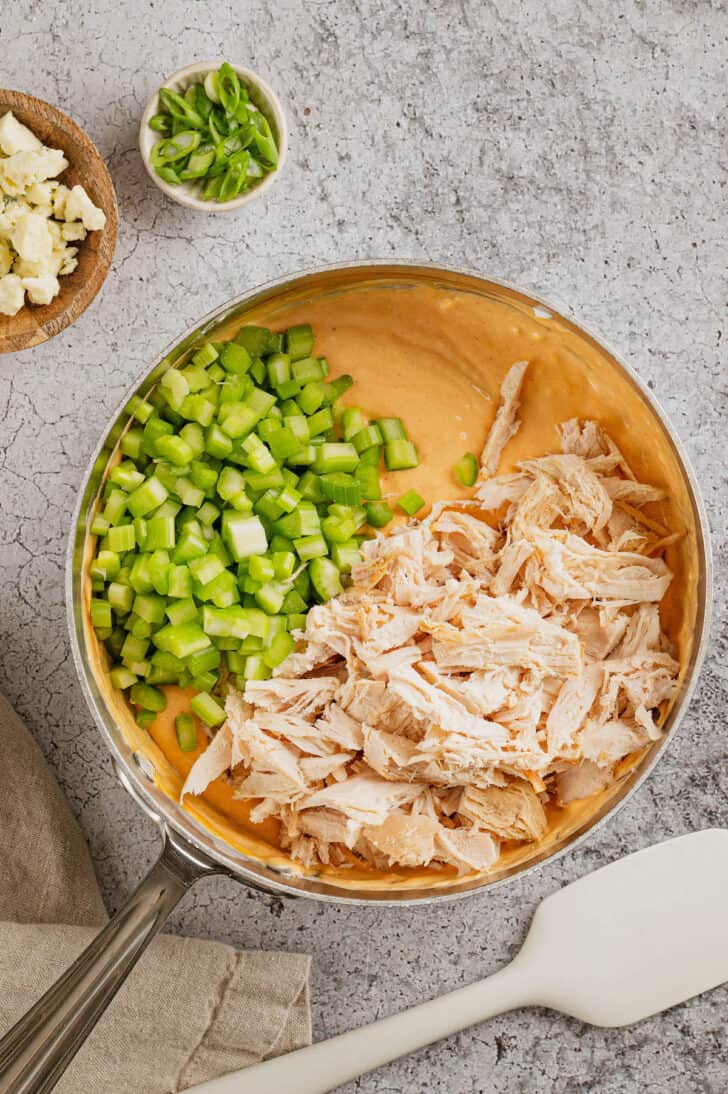 Greek yogurt buffalo chicken dip in the process of being made, with shredded chicken and chopped celery about to be stirred into the orange dip.