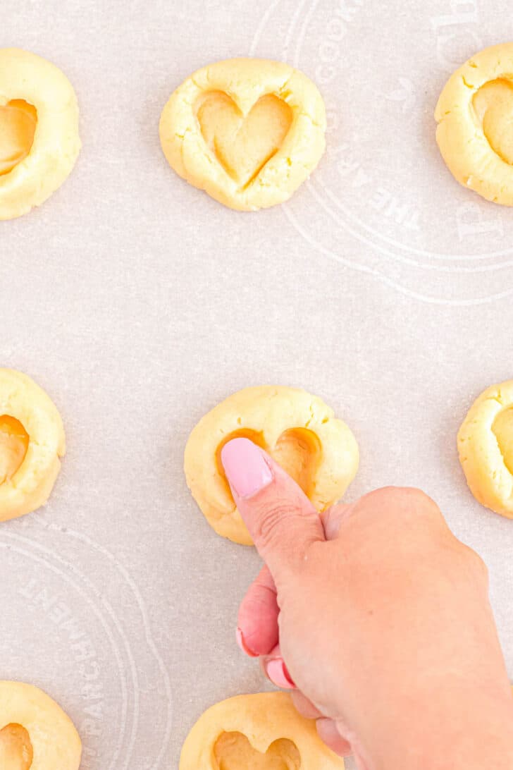 A thumb stamping a heart shape into a shortbread cookie for strawberry jam cookies.