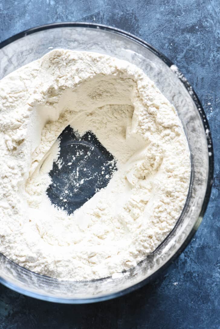 A glass bowl of flour with a well in the middle of it.