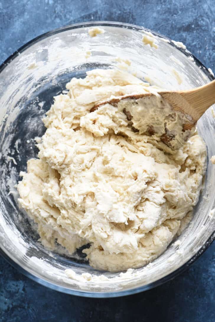 A wooden spoon stirring up a shaggy homemade pizza dough in a glass bowl.