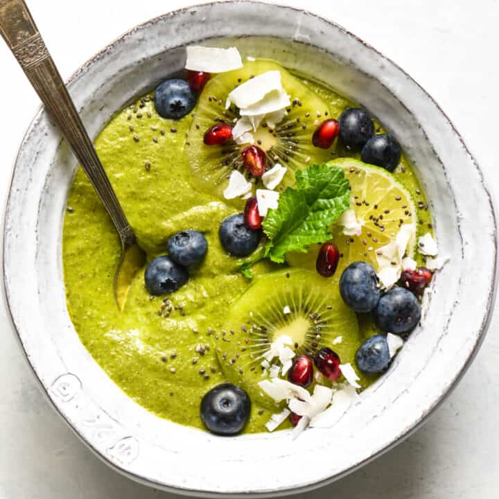 A green smoothie bowl topped with blueberries, pomegranate seeds, coconut, kiwi and a green leaf.