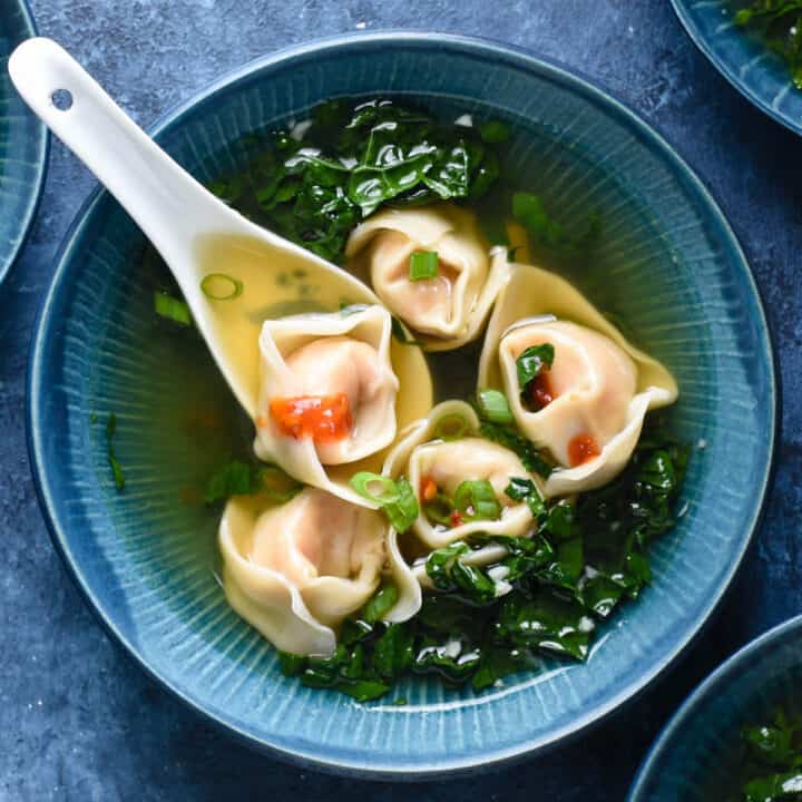 Shrimp wonton soup with kale in a blue bowl with a white soup spoon scooping up one of five wontons.