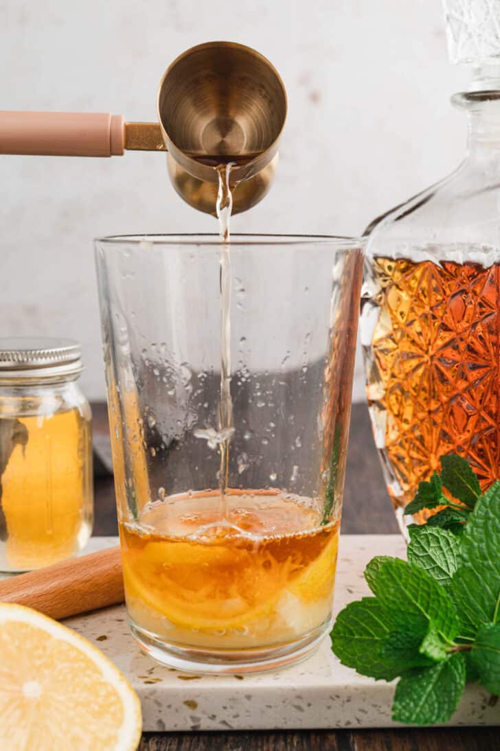 Whiskey being poured into a tall glass with crushed lemon.