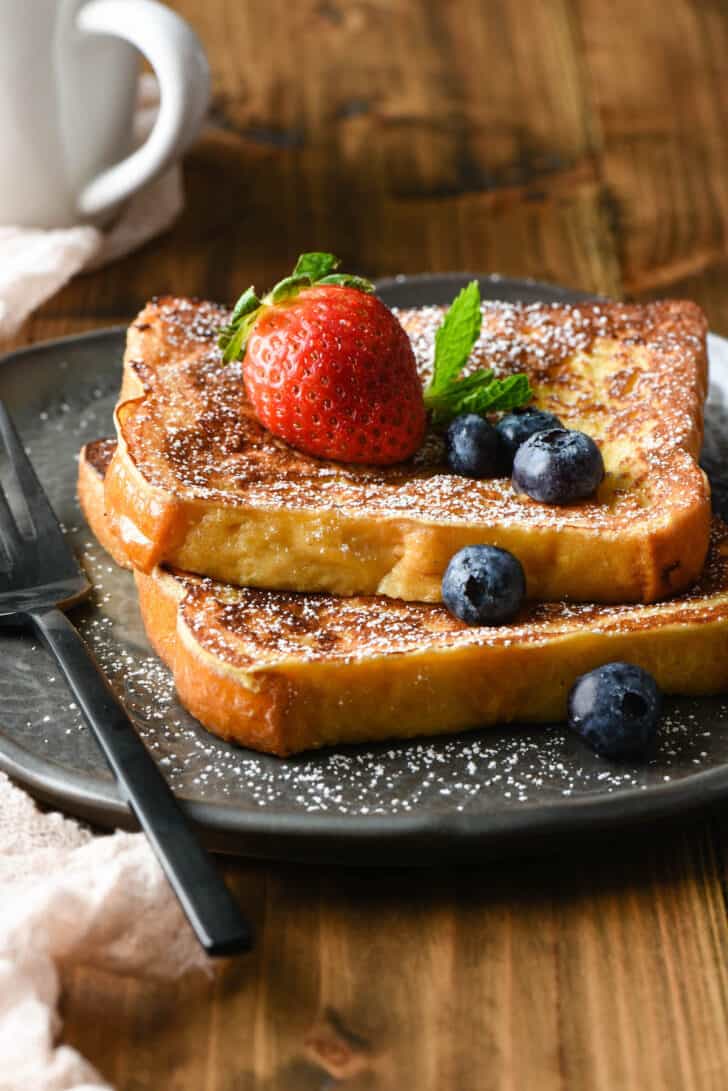 Two slices of ice cream French toast on a gray plate, topped blueberries, mint and a strawberry.