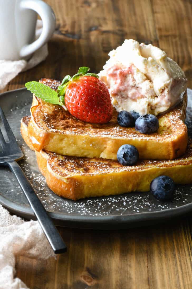 Two slices of griddled bread on a gray plate, topped blueberries, mint, a strawberry and a scoop of frozen custard.