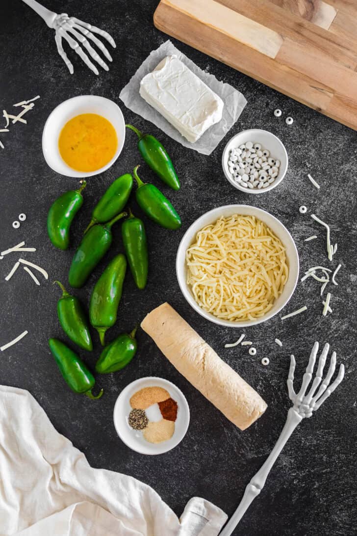 Ingredients on a dark surface, including cream cheese, cheese, green chili peppers, a roll of dough, spices and a beaten egg.