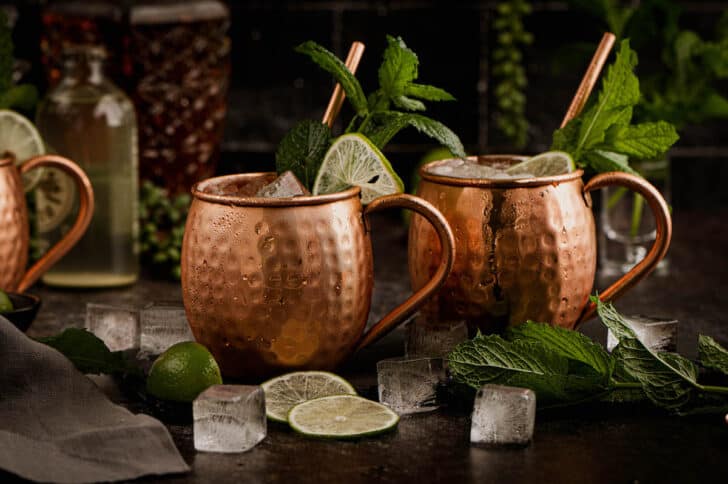 Two hammered copper mugs filled with Moscow mules with whiskey, garnished with limes and mint.