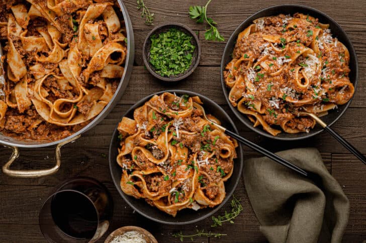 A large skillet and two black bowls on a wooden surface, all filled with noodles covered in a red meaty sauce and garnished with parsley.