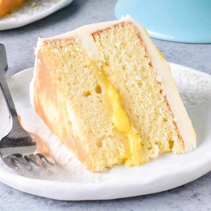 A slice of lemon curd cake with buttercream frosting on a small decorative white plate with a fork alongside.