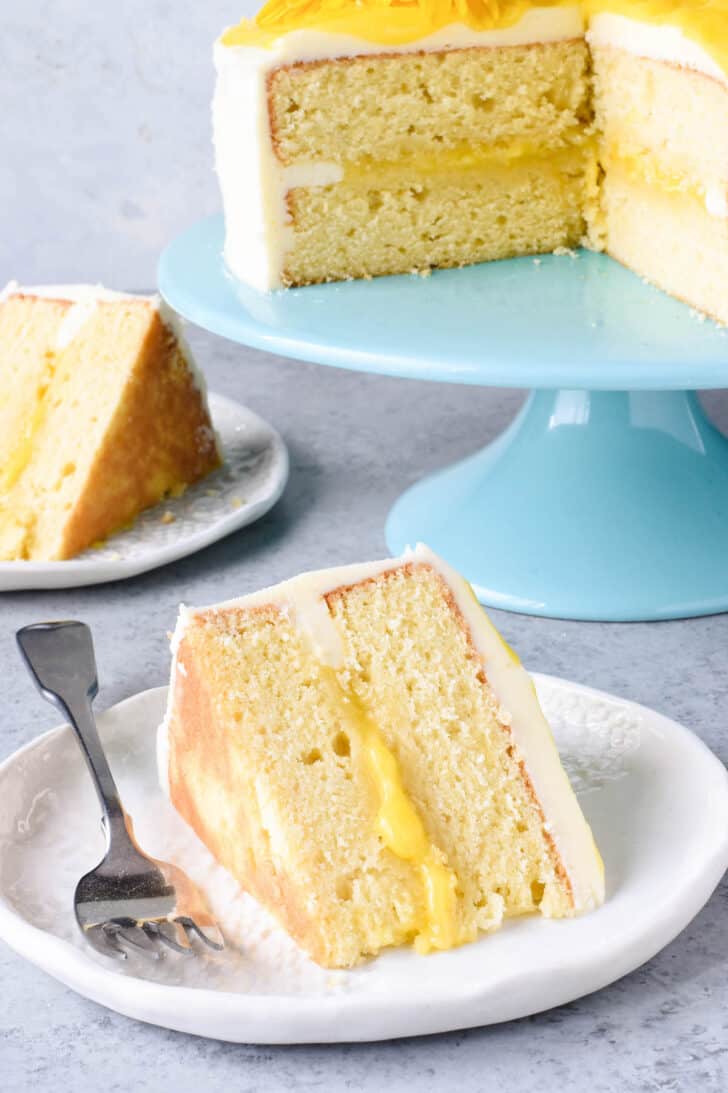 A slice of yellow cake with lemon curd filling and buttercream frosting on a small white decorative plate.