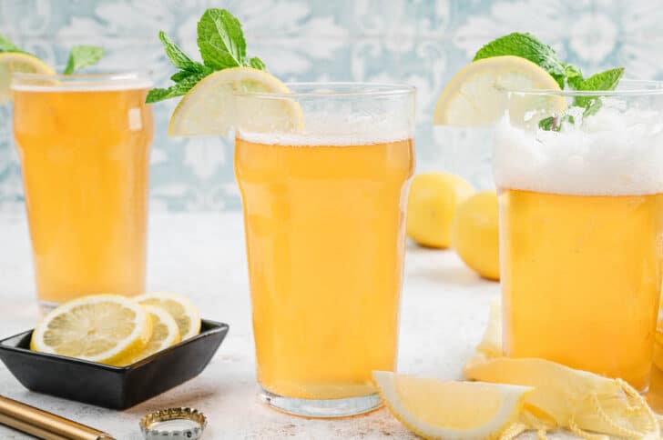 Three glasses filled with lemon shandy beer in front of light blue wallpaper.