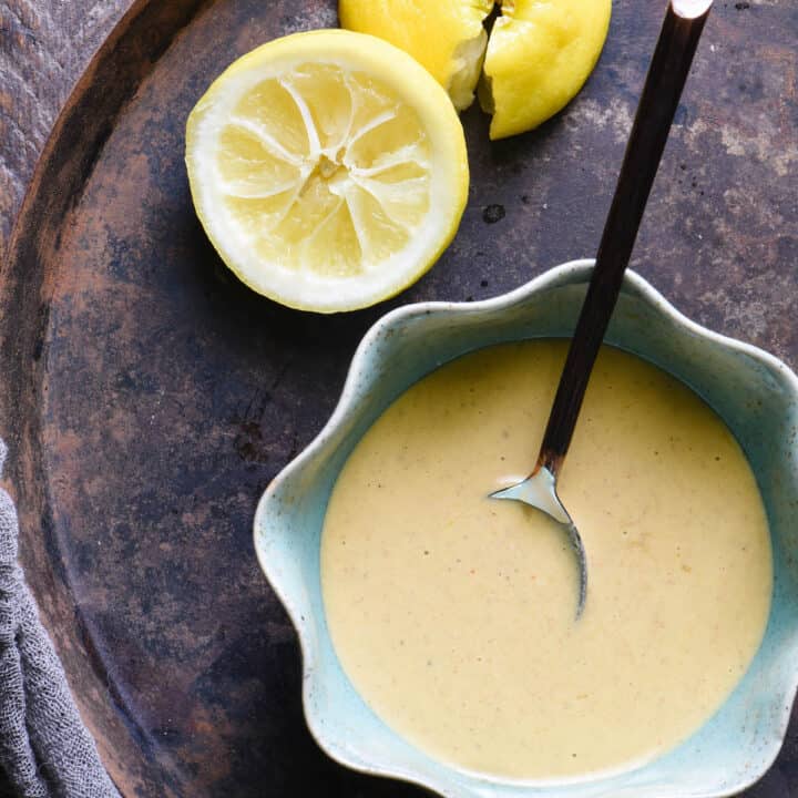 Overhead shot of bowl of tahini dressing with spoon. Lemon halves and garlic cloves on tray with dressing.
