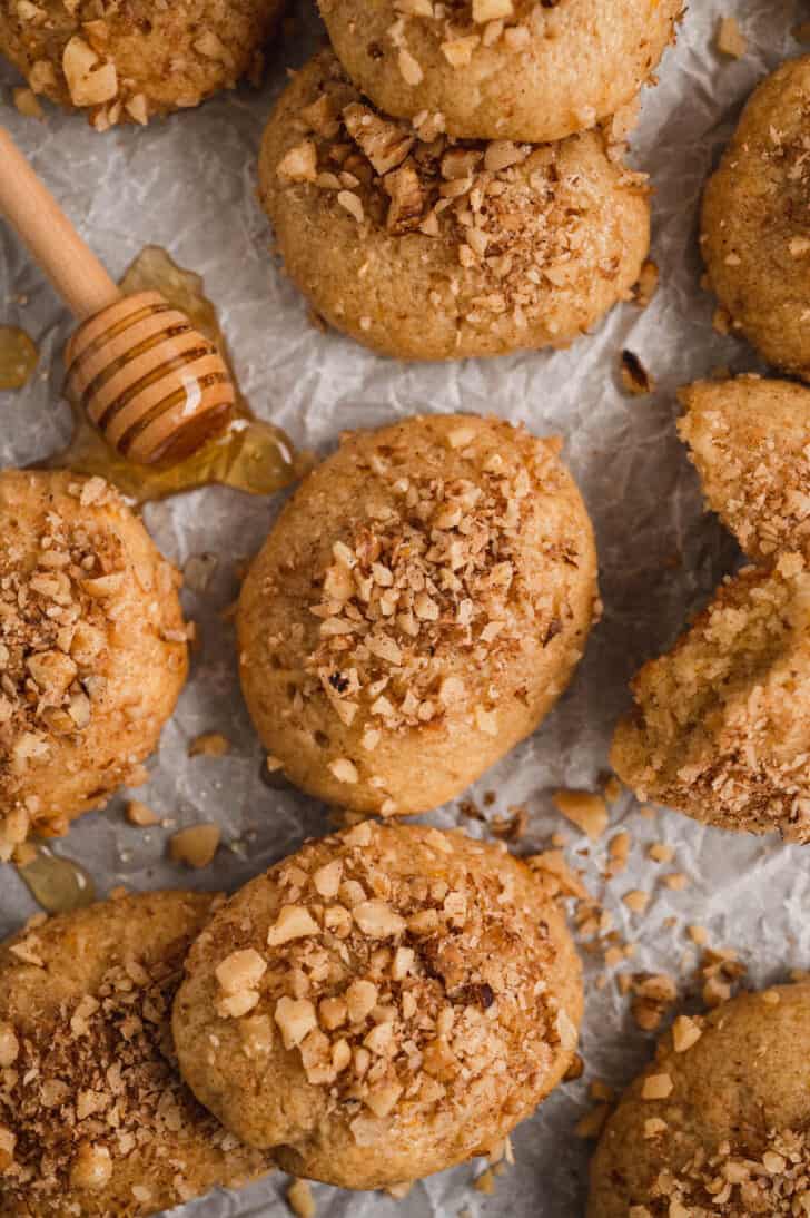 Melomakarona sprinkled with walnuts on white parchment paper with a honey dipper.