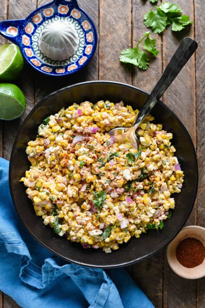 Elote salad in black bowl on wooden surface with blue napkin.