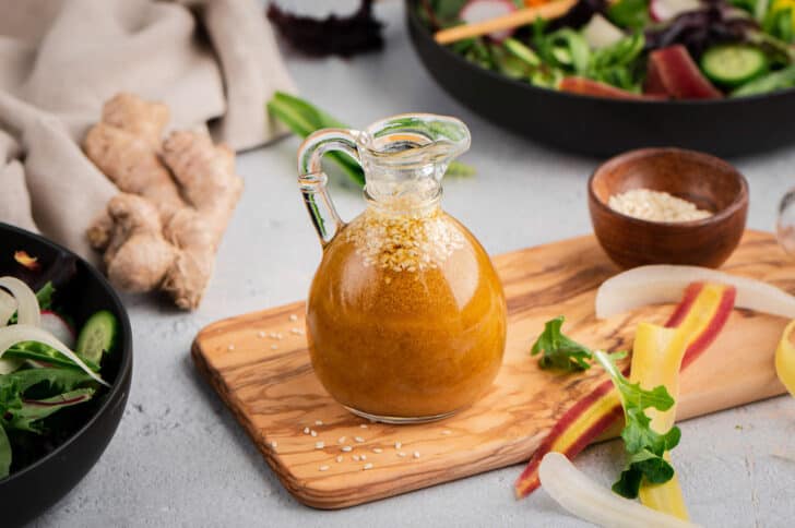 A small glass pitcher of a golden brown vinaigrette sitting on top of a small wooden cutting board, surrounded by fresh ginger and vegetables.