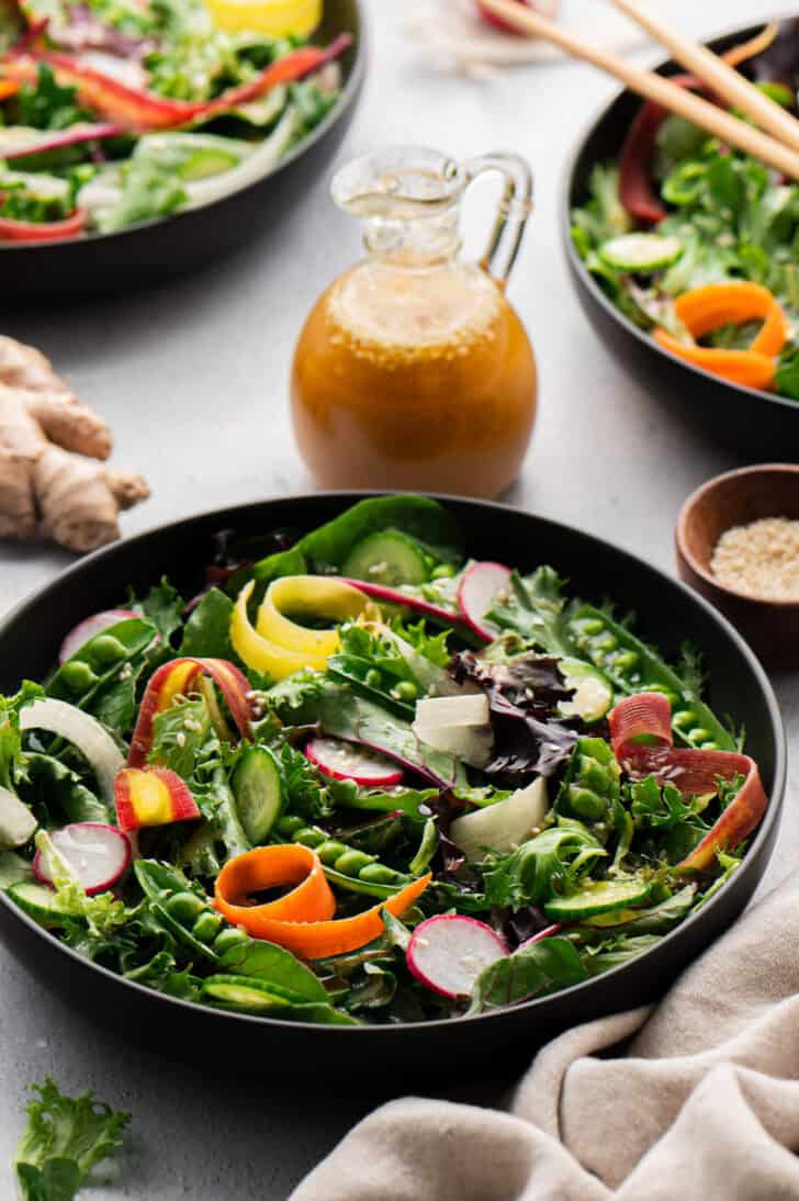 A large shallow black bowl filled with fresh vegetable salad, with a pitcher of miso ginger dressing in the background.