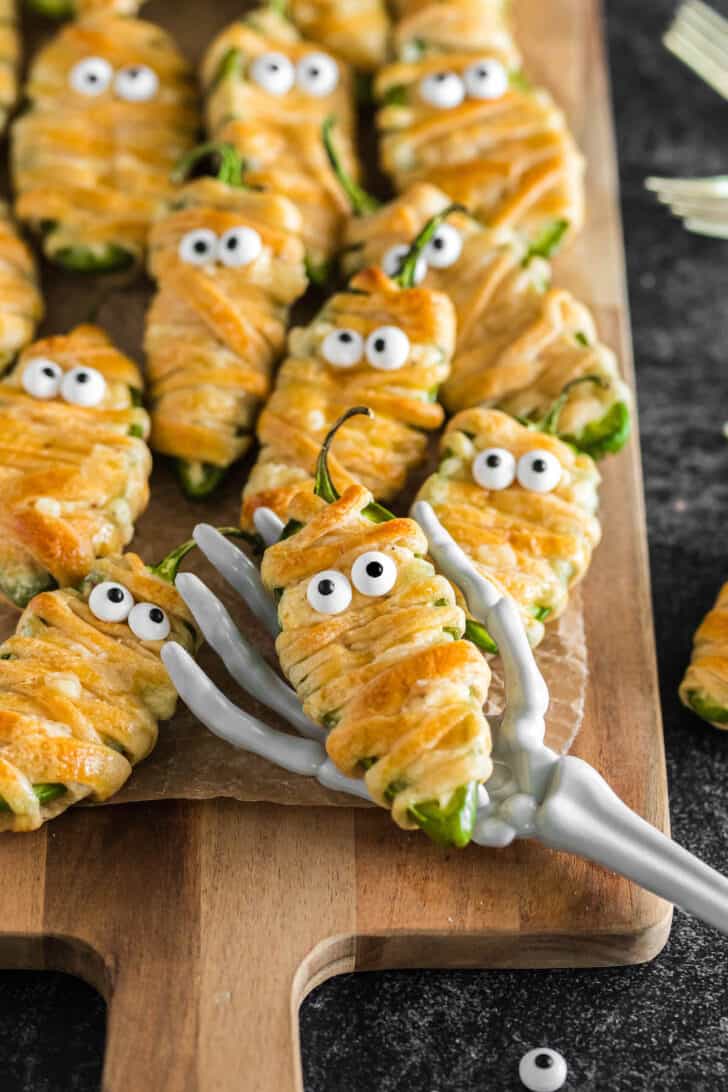 A toy skeleton hand holding one of the mummy jalapeno poppers on display on a cutting board.