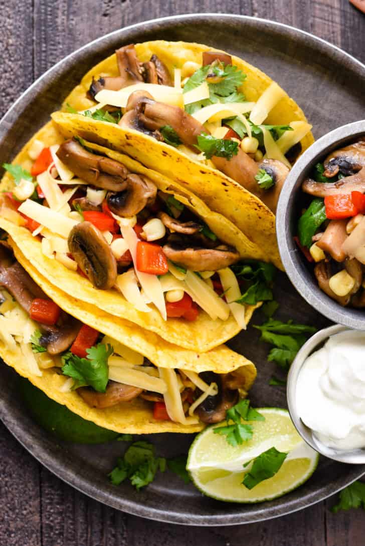 A round tray filled with three mushroom tacos made in corn tortillas, a lime wedge, and small bowls of mushroom filling and sour cream.