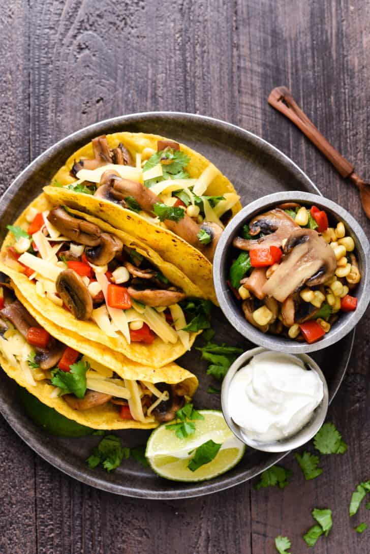 A round tray filled with three mushroom tacos made in corn tortillas, a lime wedge, and small bowls of mushroom filling and sour cream.