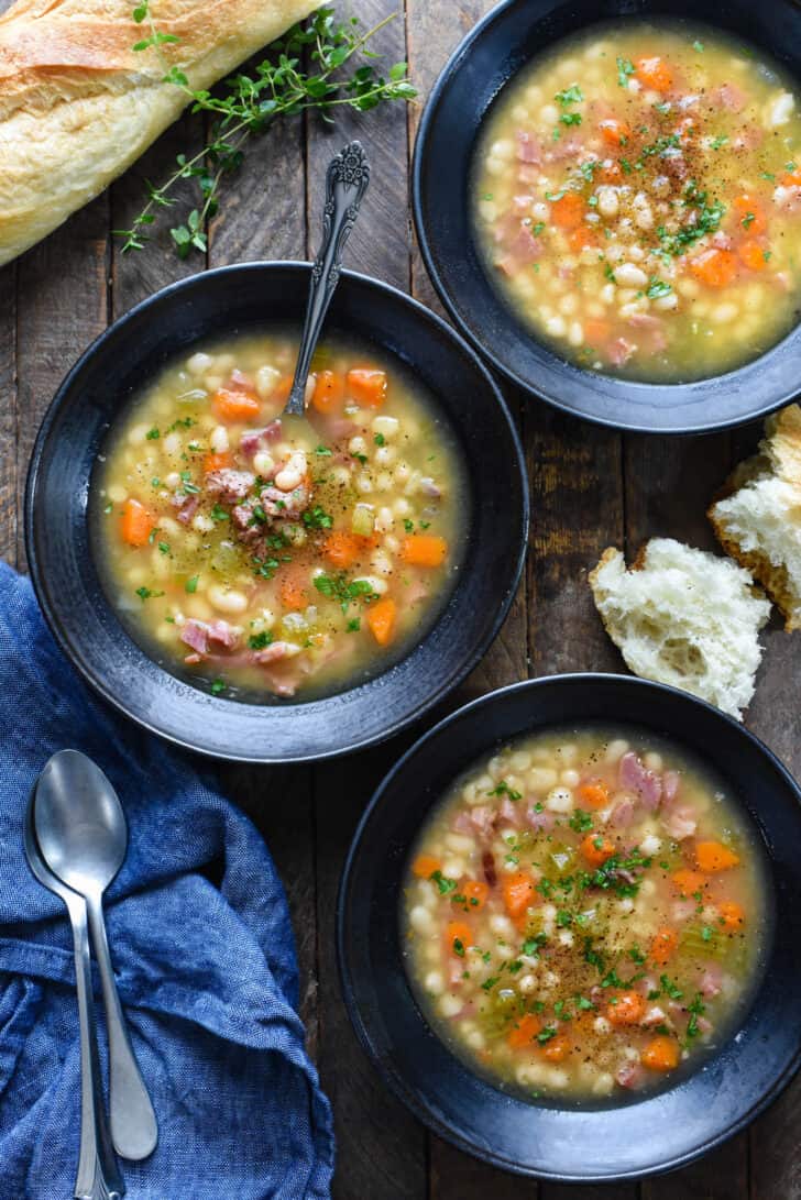 Three black bowls filled with old fashioned ham and bean soup on a rustic wooden tabletop.