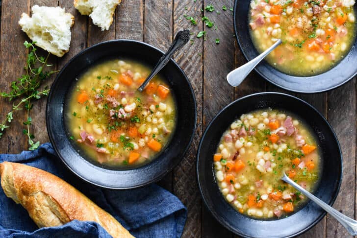 Three black bowls filled with homemade ham and bean soup on a rustic wooden background, with bread and herbs decorating the scene.