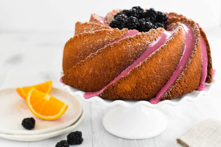 A spiral shaped cake decorated with purple icing and blackberries on a fluted white ceramic cake stand.