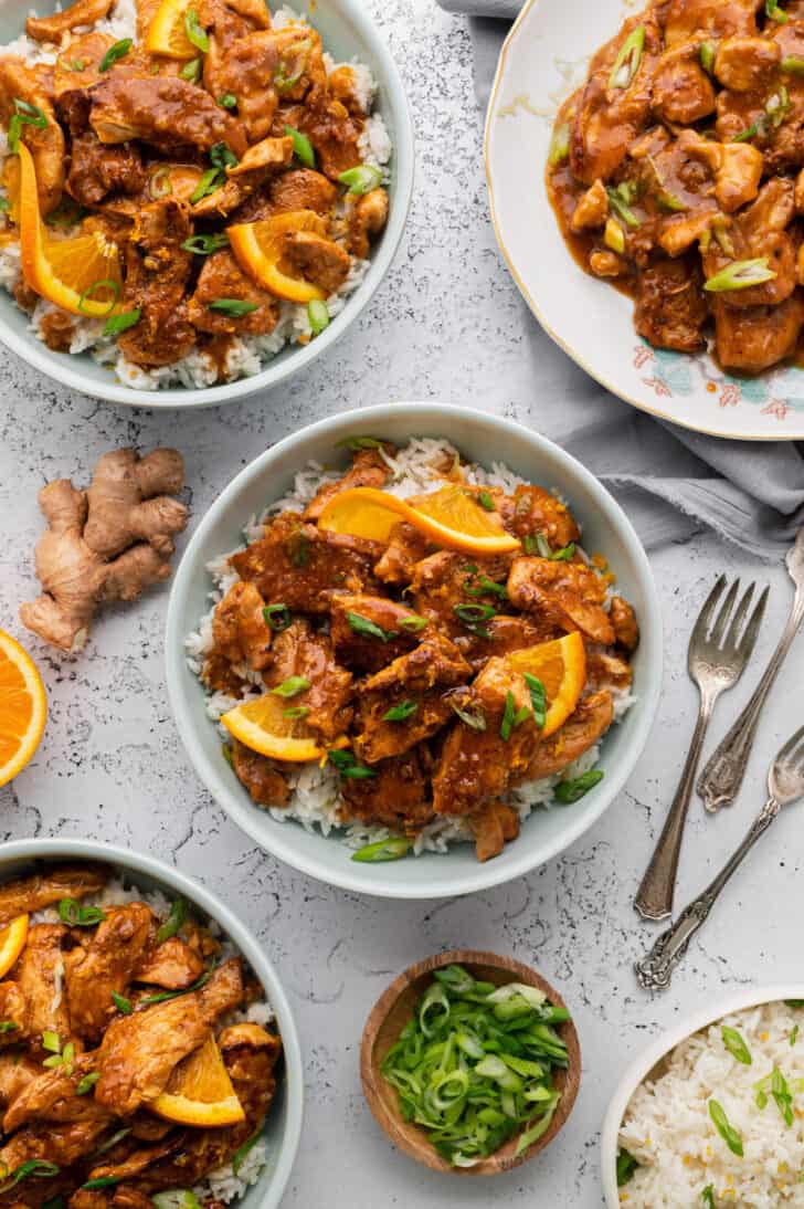 Several bowls and plates of orange ginger chicken served over rice on a light surface.