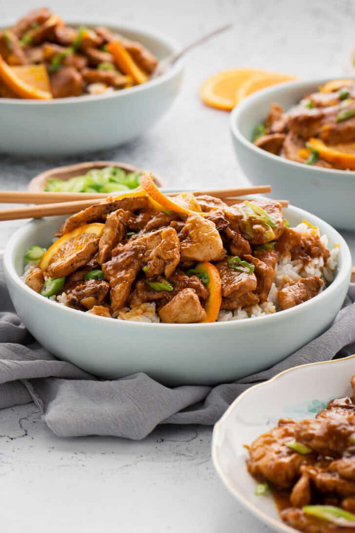 White bowls filled with rice and orange ginger chicken with a set of chopsticks taking out a bite.