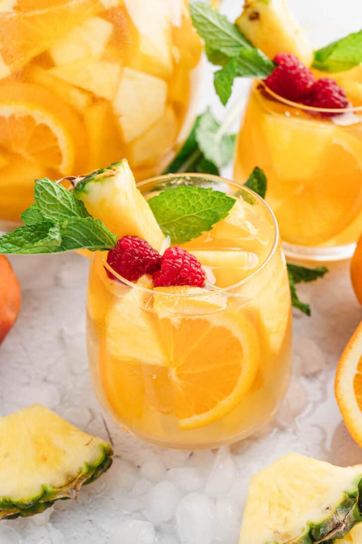 Glasses of pineapple sangria with mango over ice, garnished with raspberries and fresh mint sprigs.