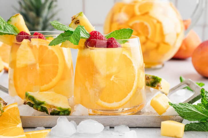 Glasses of pineapple sangria with mango over ice, garnished with raspberries and fresh mint sprigs.
