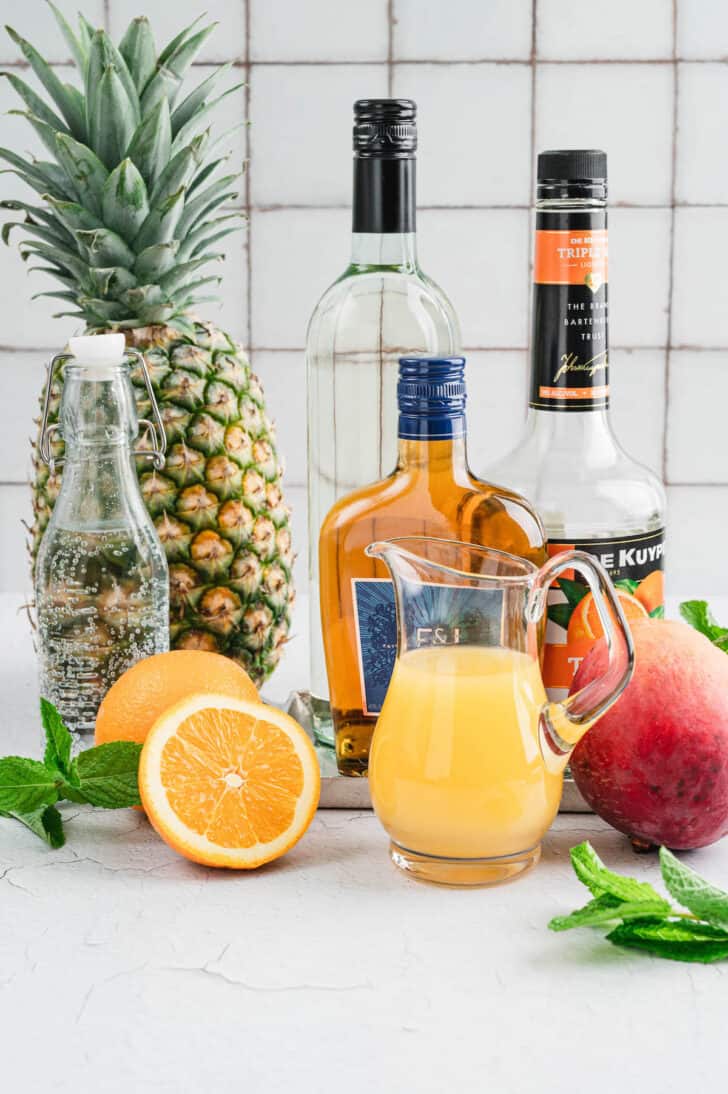 Ingredients for a cocktail arranged on a white counter, including bottles of brandy, triple sec, sparkling water and citrus fruits.