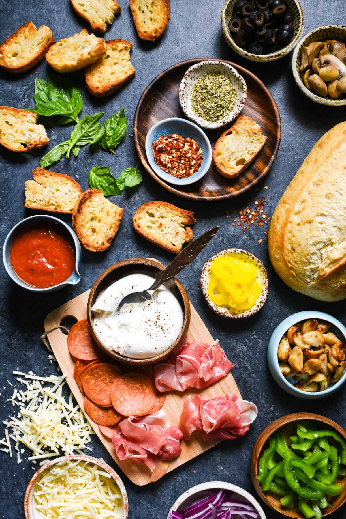 Pizza Crostini Bar - Put out some toasted bread and a big spread of pizza toppings, and let everybody make their own appetizer. So fun for parties! | foxeslovelemons.com A dark surface filled with the toppings for a crostini pizza bar, including toasted bread, meats, cheese, vegetables and herbs and spices.