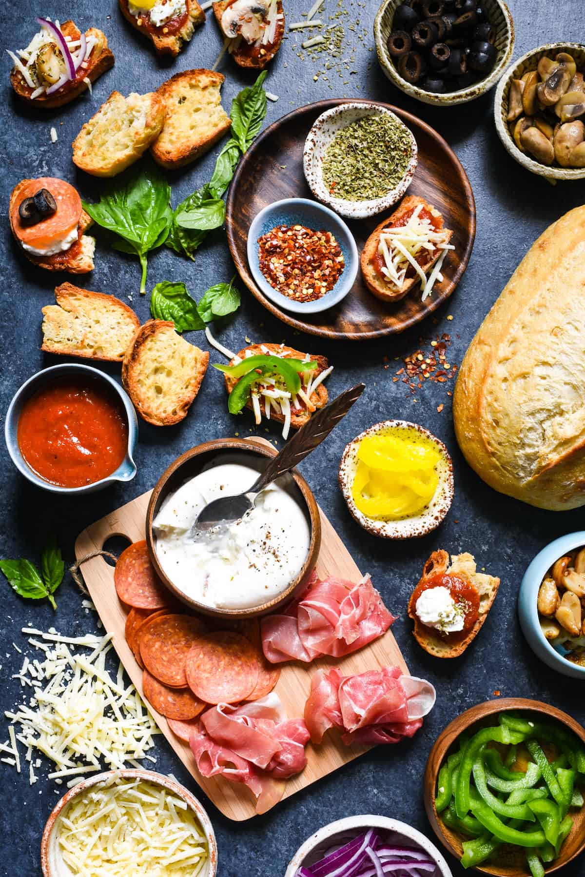 Pizza Crostini Bar - Put out some toasted bread and a big spread of pizza toppings, and let everybody make their own appetizer. So fun for parties! | foxeslovelemons.com A dark surface filled with the toppings for a crostini pizza bar, including toasted bread, meats, cheese, vegetables and herbs and spices.