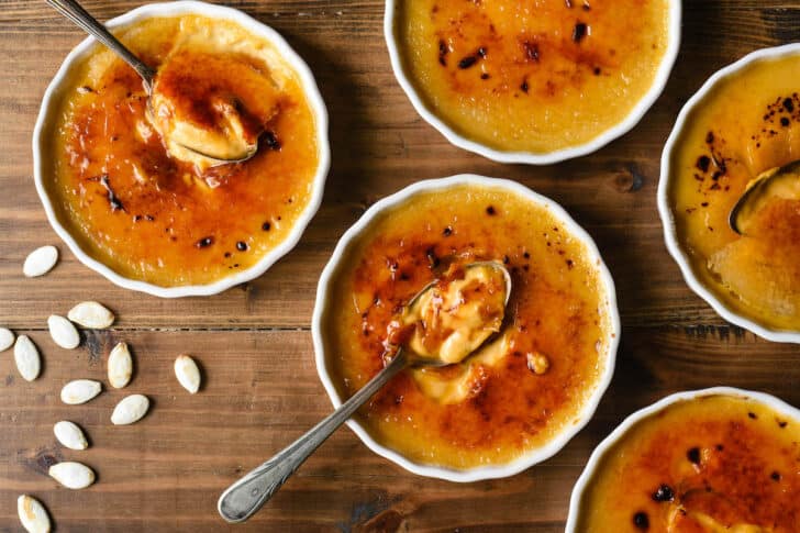 Shallow white ramekins filled with pumpkin spice creme brulee on a wooden tabletop, with pumpkin seeds and spoons garnishing the scene.