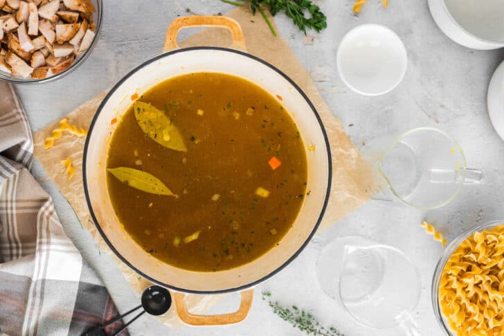 A white Dutch oven with a broth and vegetable in it, with two Bay leaves floating on top.