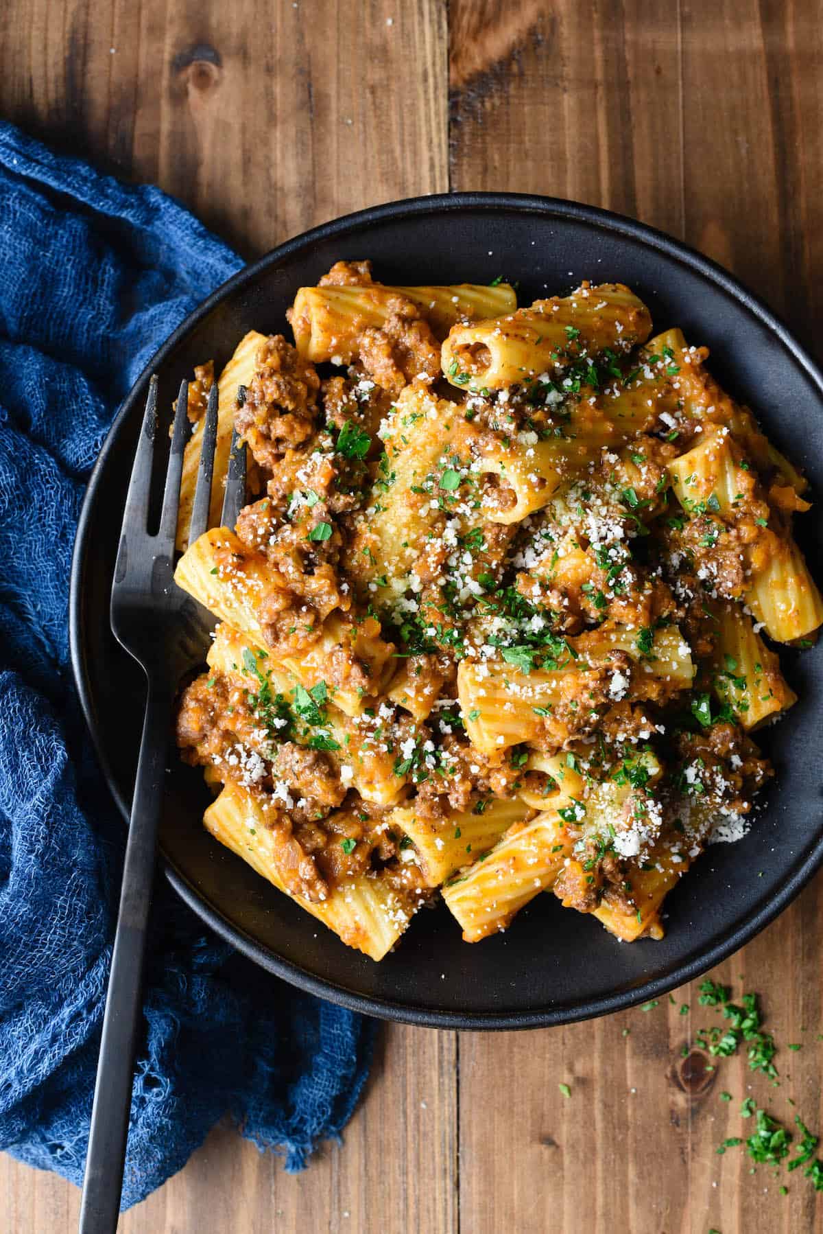 A bowl of this Rigatoni Bolognese will hit the spot whenever you have a pasta craving. This easy but elegant meat sauce recipe will give you a restaurant quality dish right at home. | foxeslovelemons.com Overhead photo of rigatoni with meat sauce in black bowl, with black spoon and blue linen alongside.
