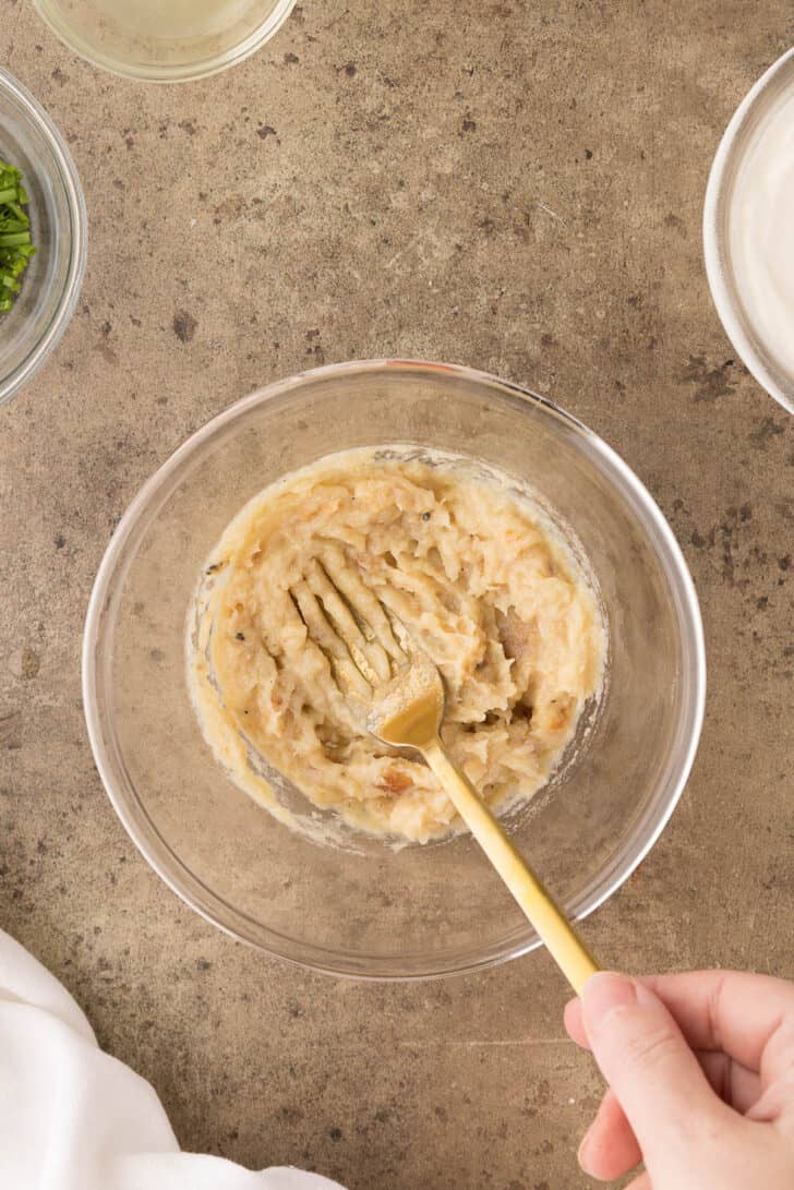 A glass bowl filled with mashed roasted garlic, with a gold fork mashing the mixture.