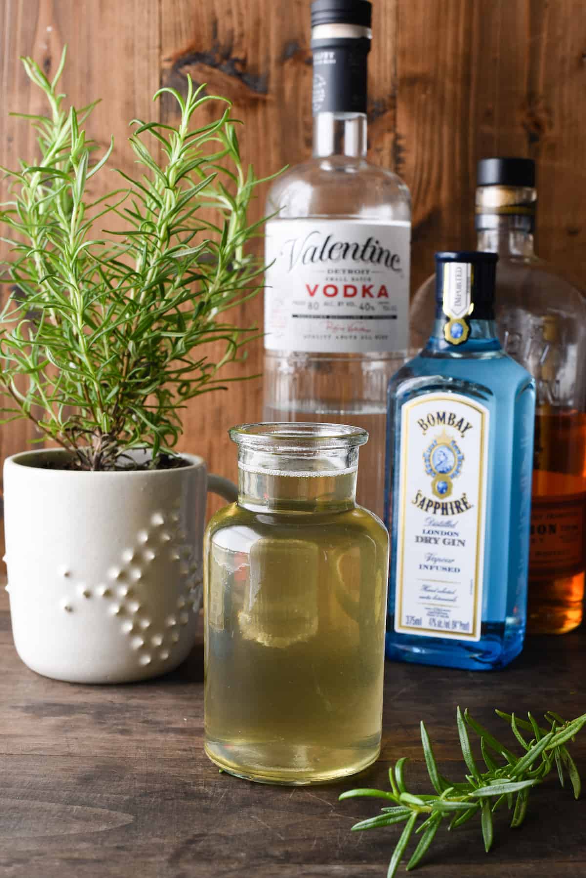 This homemade Rosemary Simple Syrup will make you feel like a fancy bartender, right in your own kitchen. Whip up a batch of this, and use it for whisky, gin, vodka or mocktails. | foxeslovelemons.com Small bottle of rosemary simple syrup with liquor bottles and rosemary plant in background.